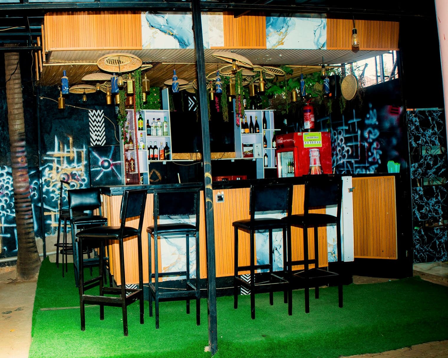 The outdoor bar: black leather stools on green turf, a timber-clad counter, bottles hung from woven baskets and graffiti murals behind